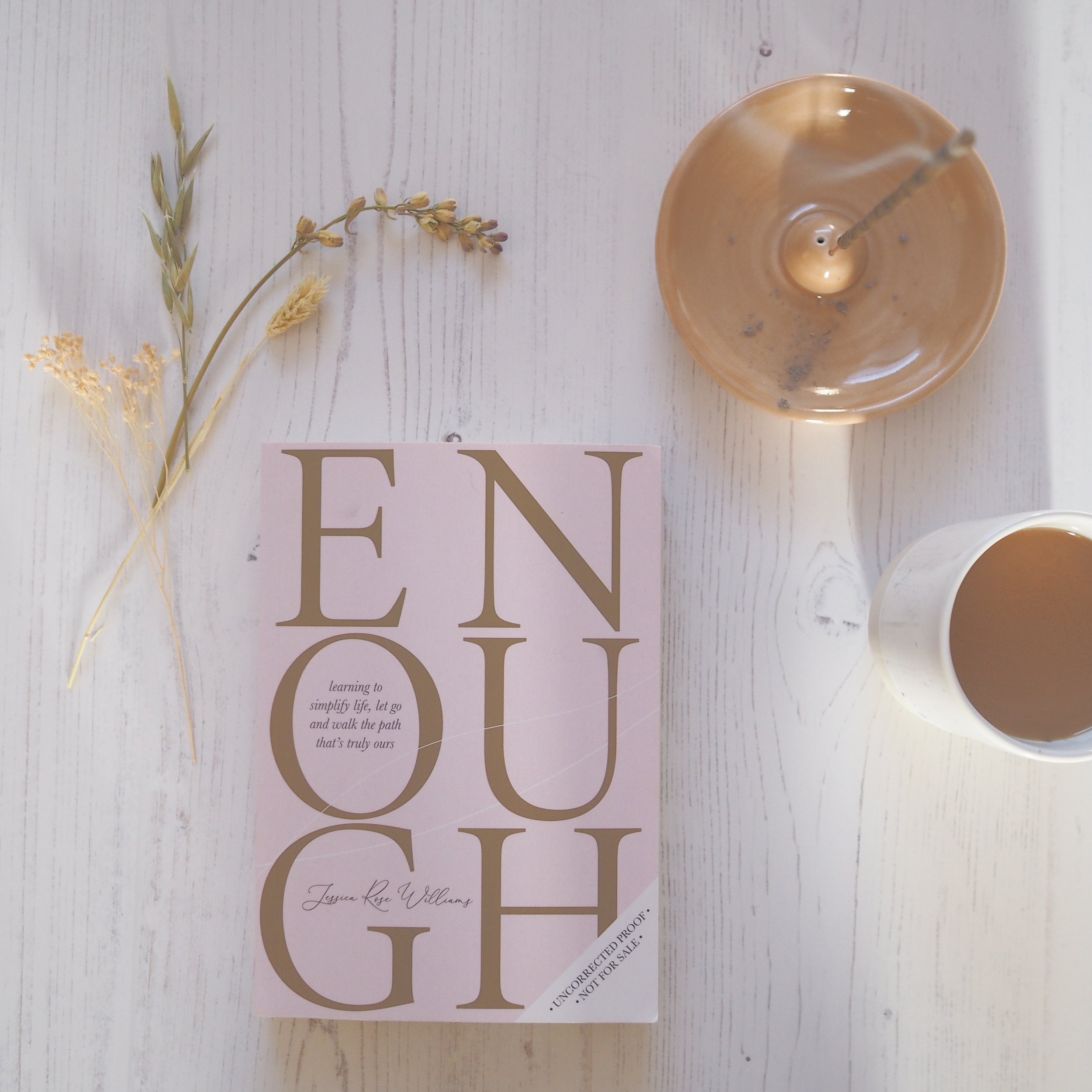 Book called Enough on white surface, with dried flowers, incense and cup of tea.