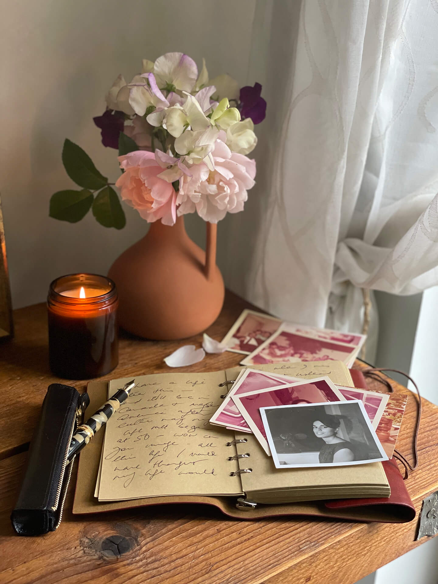 Open writing journal on a desk with old photographs next to a vase of flowers and a candle.