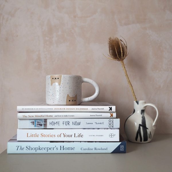 Stack of lifestyle books on a desk, with a mug on top and a vase with a dried seedhead