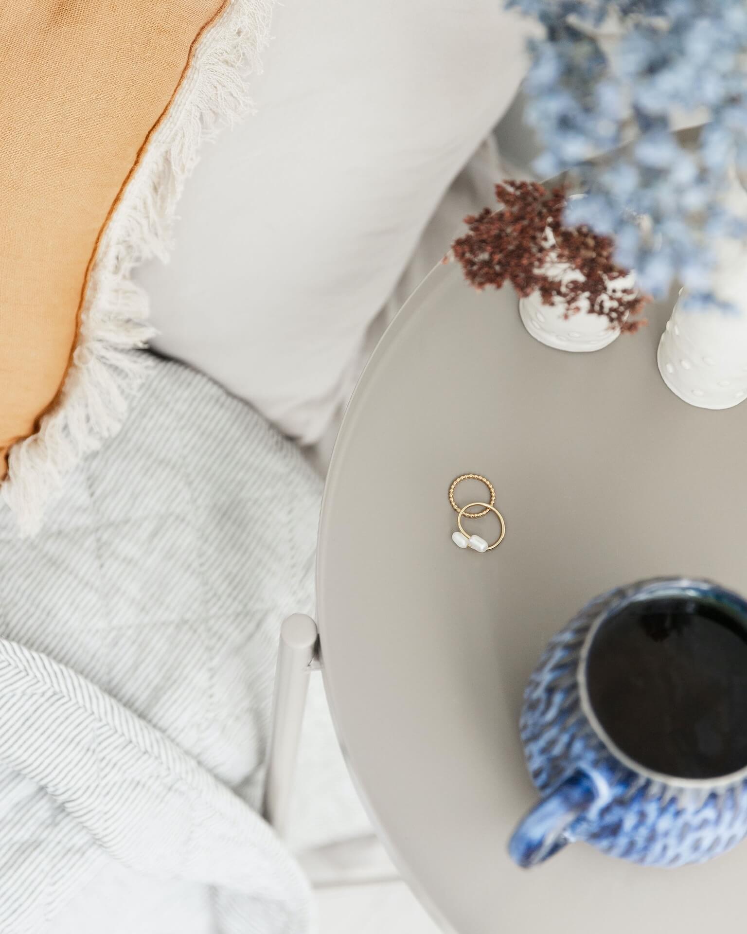 bedside table with jewellery, blue mug and Ochre cushion on a bed and other items from lifestyle store Tusk