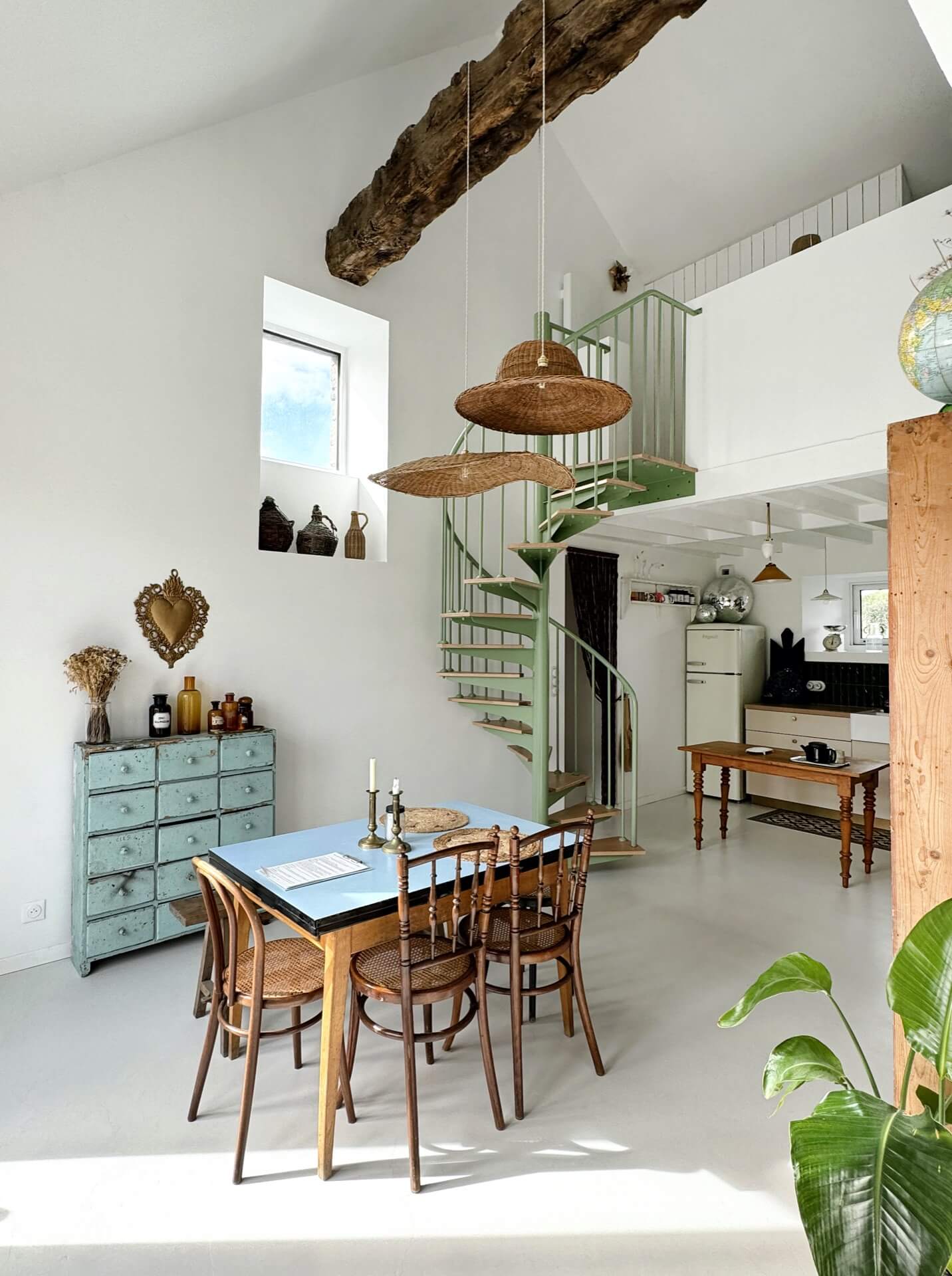 an open plan kitchen and dining area in an old French barn conversion, with a mint green spiral staircase and lots of vintage furniture and objects decorating the space. 